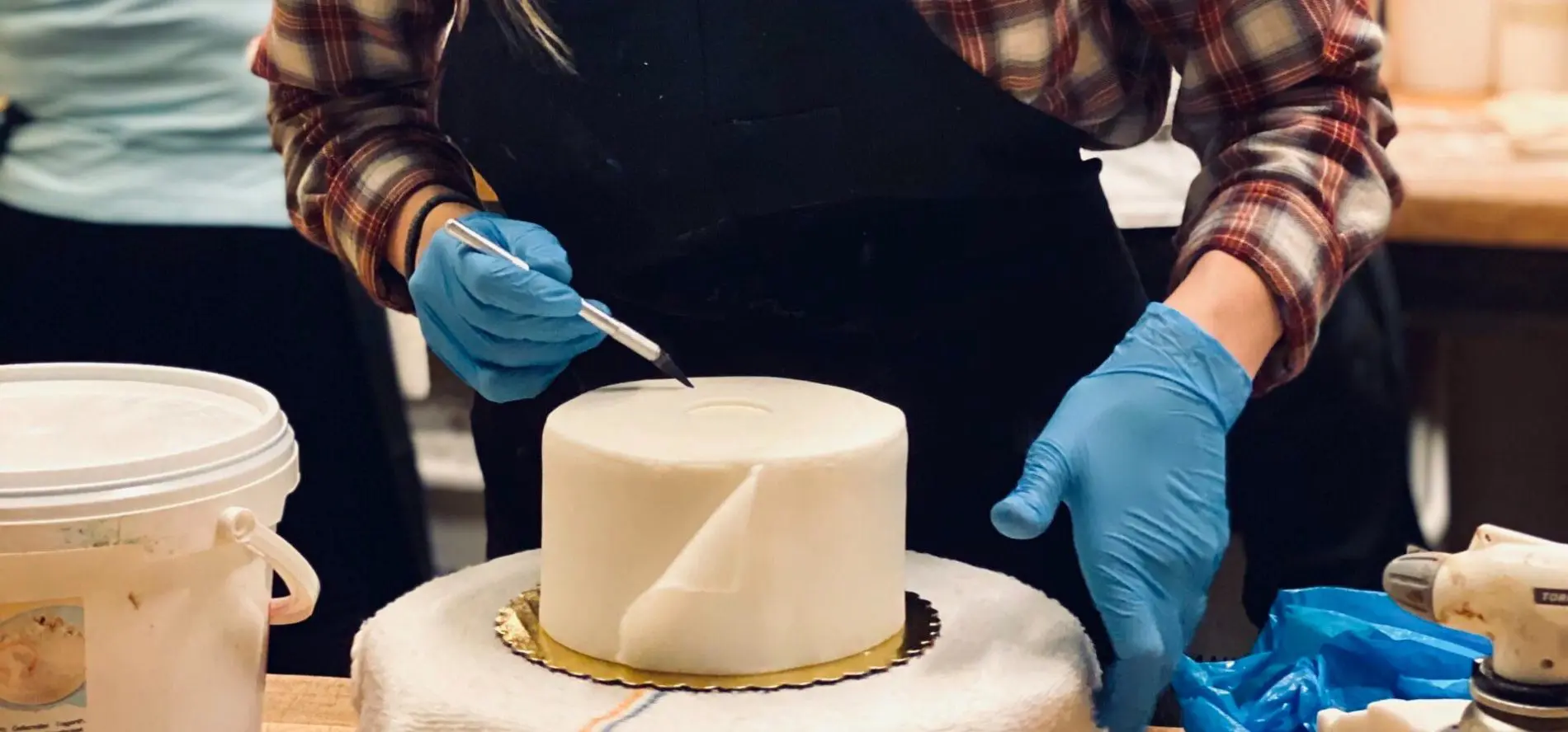 Seattle Catering A person wearing a plaid shirt and blue gloves is carefully decorating a round, white cake on a turntable using a tool, showcasing the meticulous skills celebrated in jobs at Lisa Dupar Catering.