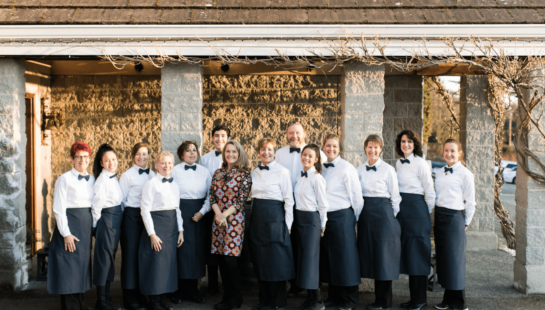 Seattle Catering A group of thirteen people, mostly dressed in white shirts, black bow ties, and black aprons, stands together in front of a building, with one person in a patterned dress in the center. They represent the dedicated team behind jobs at Lisa Dupar Catering.