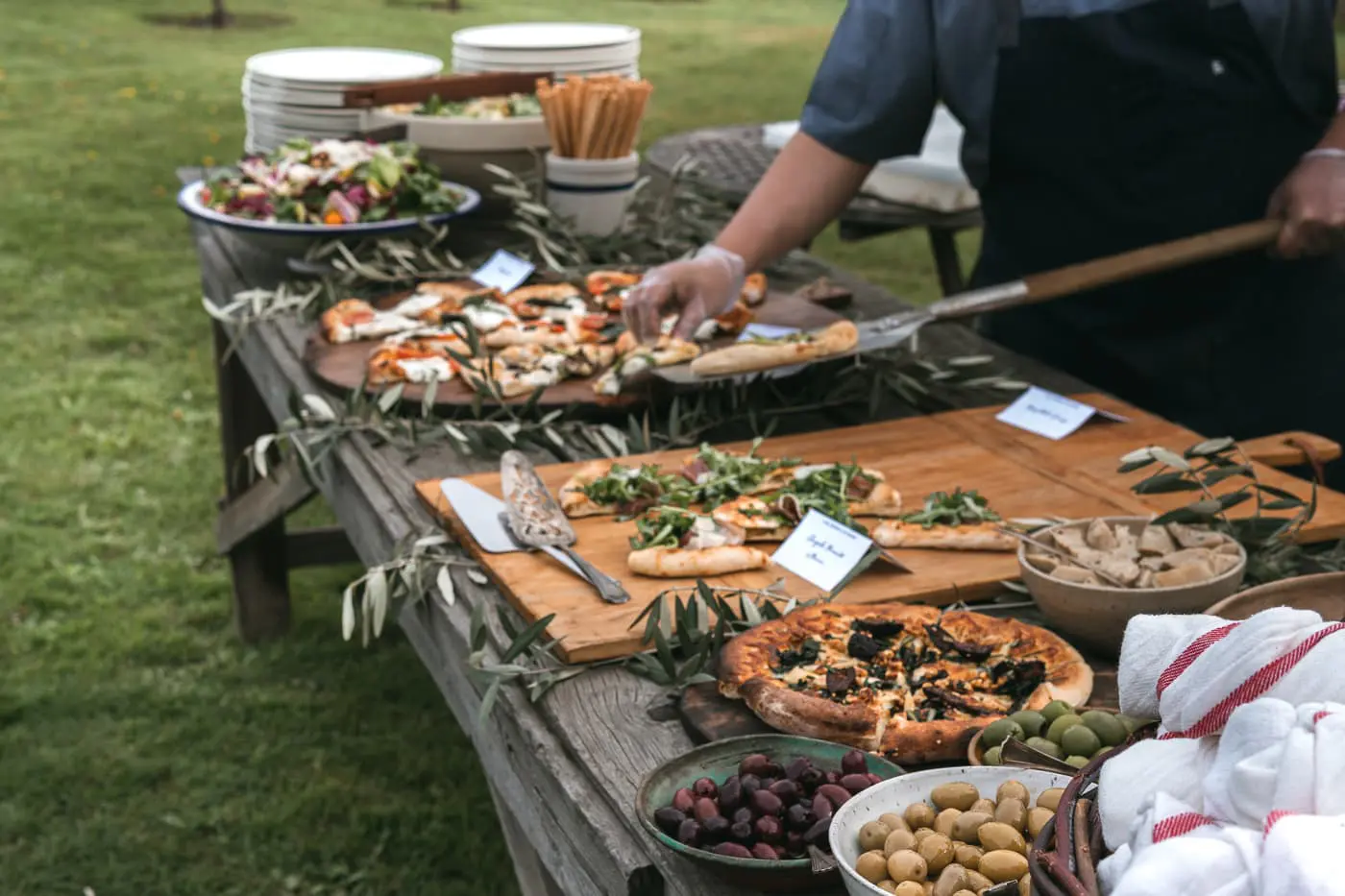 Seattle Catering A person is preparing assorted pizzas on a wooden table outdoors, creating a delightful catering menu. Other foods, such as salad, breadsticks, and bowls of olives, are also on the table. Plates and serving utensils are visible in the background.