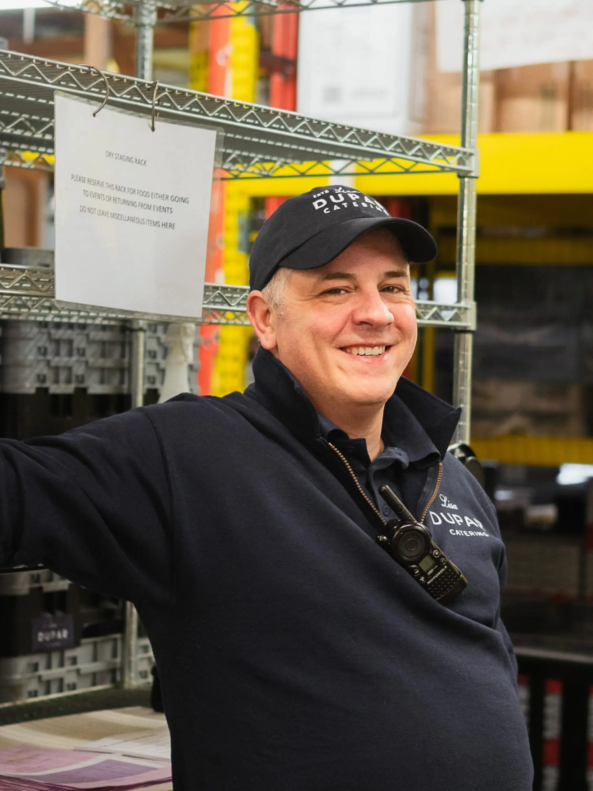 Seattle Catering A man in a warehouse, wearing a cap and nametag, standing in front of metal shelves with a sign in the background.