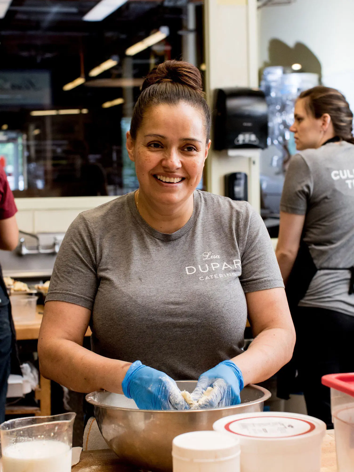 Seattle Catering A woman wearing a gray "Lisa Dupar Café" shirt and blue gloves is mixing ingredients in a bowl in a kitchen, smiling. Another person is working in the background.