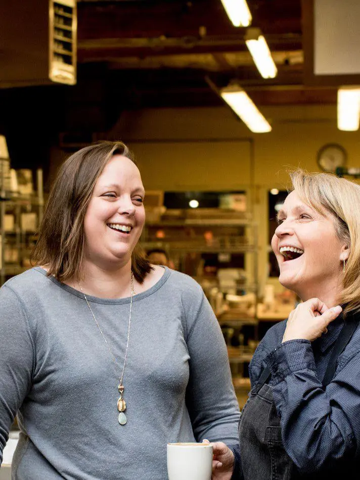 Seattle Catering Two women are standing in a kitchen, laughing and enjoying a conversation. One is holding a white mug. The background shows various kitchen items and bright lighting fixtures.