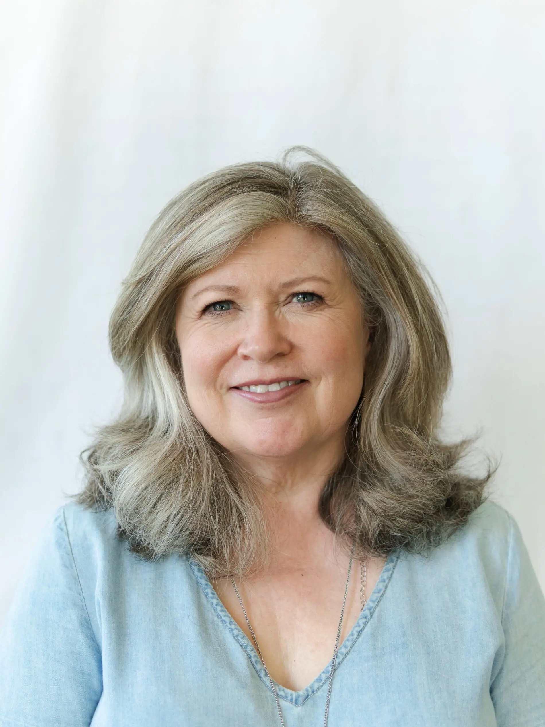 Seattle Catering A woman with grey hair wearing a light blue shirt smiles while standing against a neutral background.