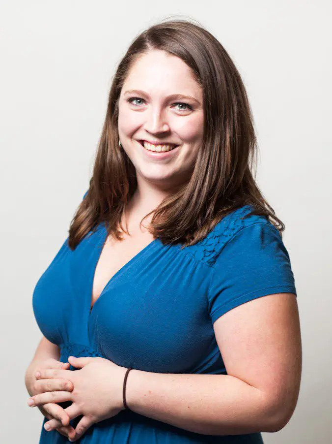 Seattle Catering A woman with long brown hair wearing a blue dress smiles at the camera with her hands clasped.