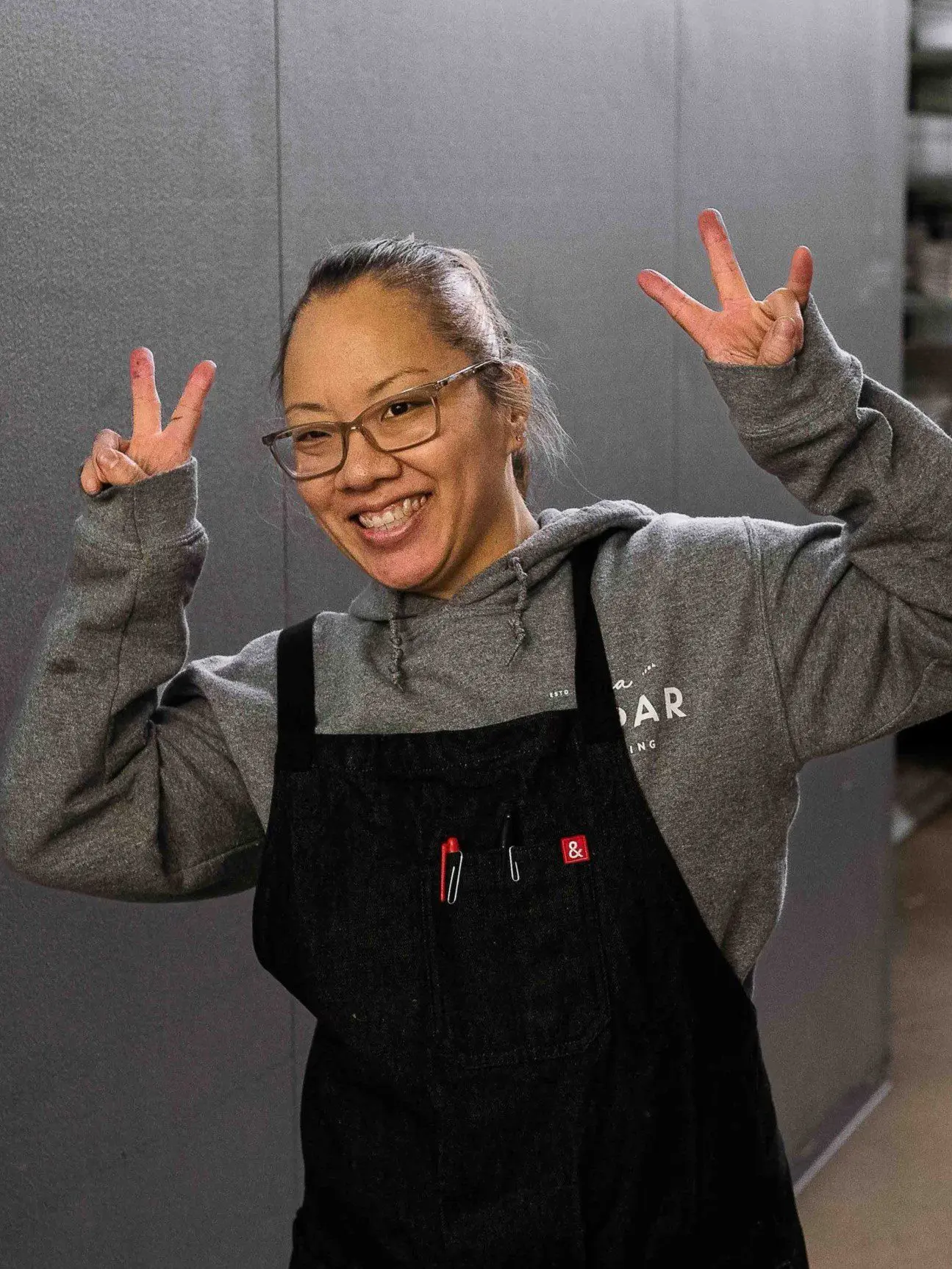 Seattle Catering A person wearing glasses, a gray hoodie, and a black apron smiles and makes a peace sign with both hands indoors.