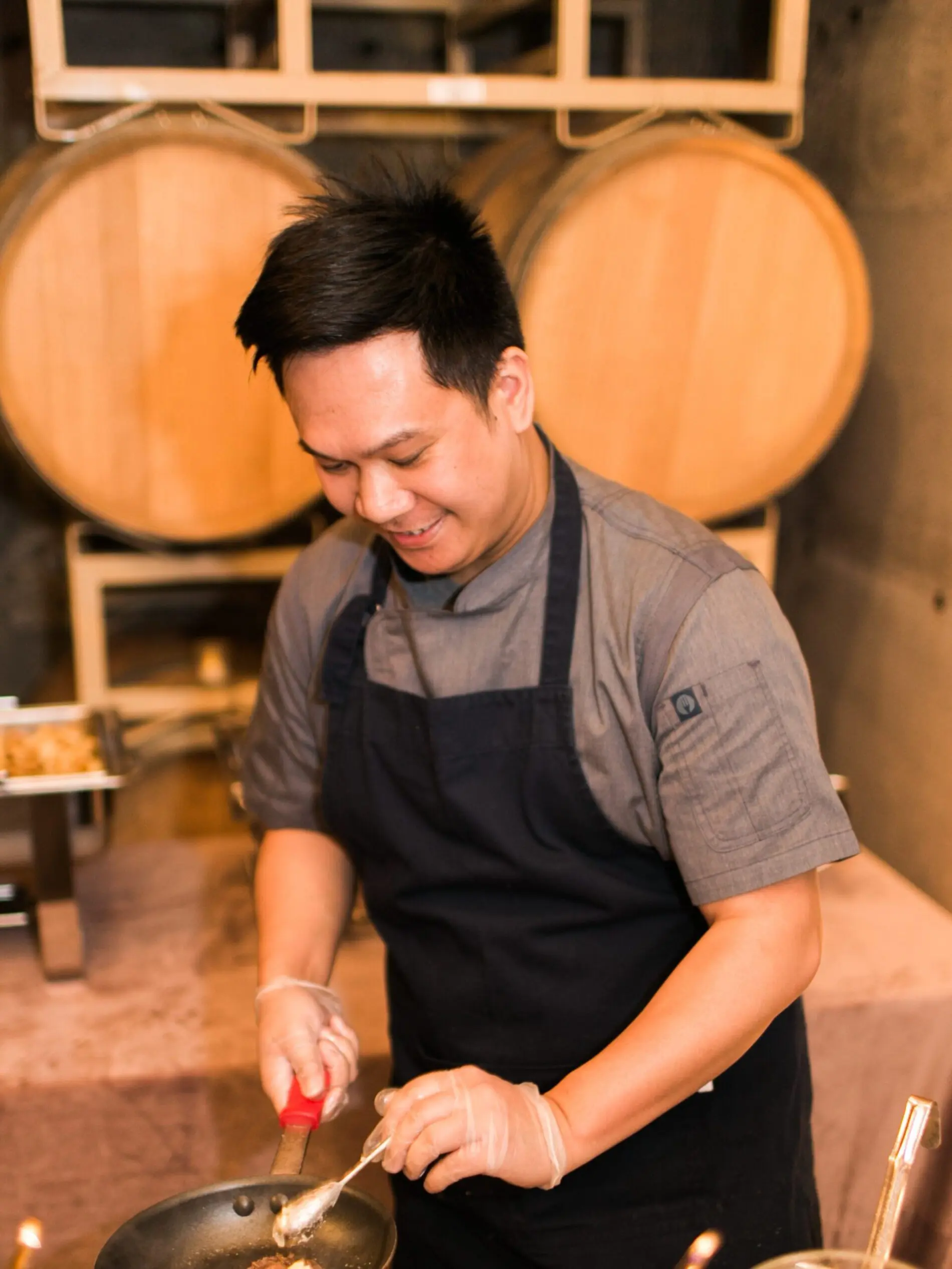 Seattle Catering A chef wearing a dark apron and gloves cooks food in a frying pan, with large wooden barrels in the background.