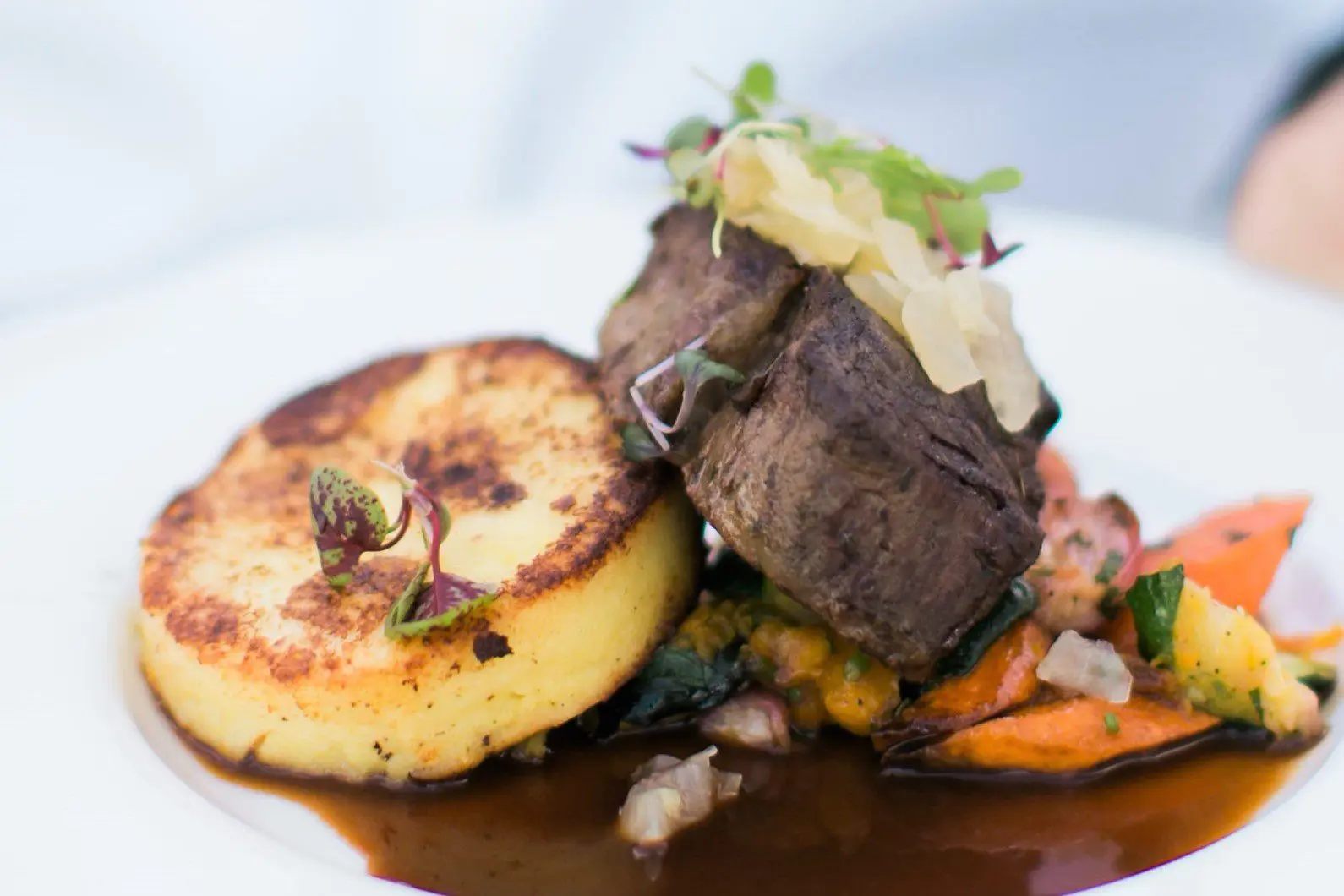 Seattle Catering A plated dish featuring a seared beef steak topped with microgreens, accompanied by a grilled polenta cake and roasted vegetables, all served on a bed of rich brown sauce.