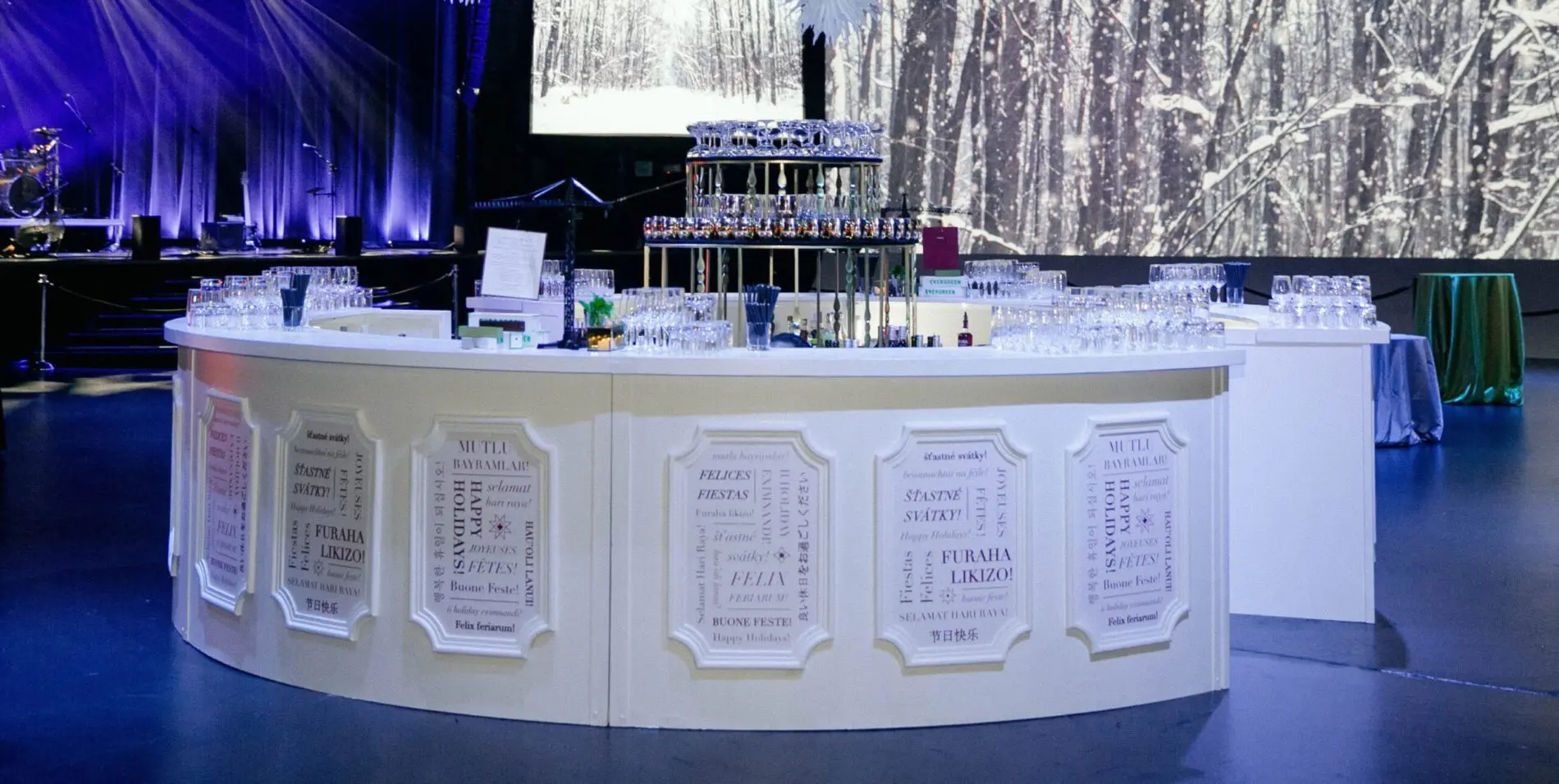 Seattle Catering A semi-circular white bar setup with various drinks on a counter, provided by top event vendors in Seattle. Glasses are neatly arranged, and the background features a snowy forest scene projection along with stage equipment.