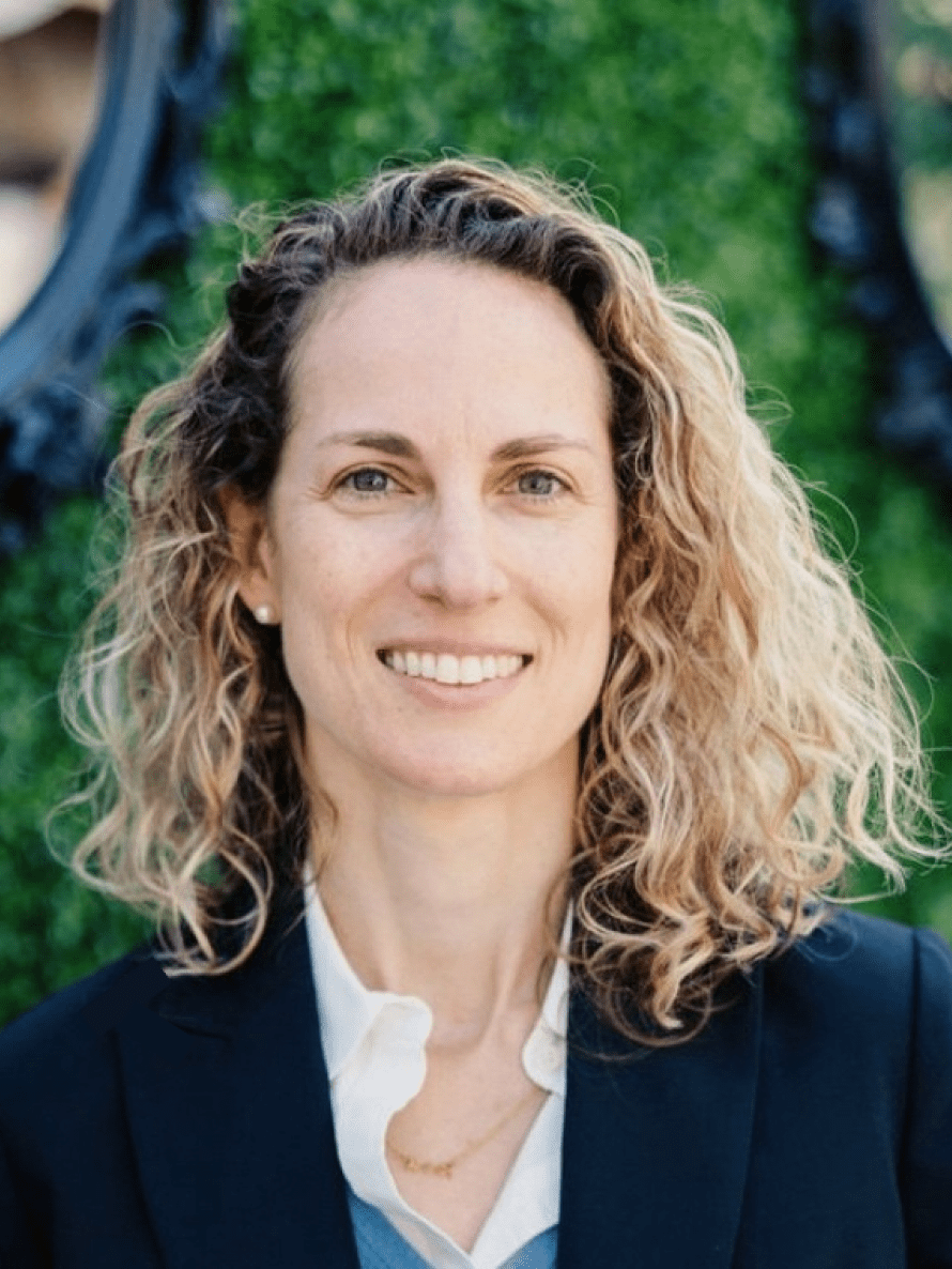 Seattle Catering A woman with curly blond hair smiles at the camera, wearing a navy blazer and white blouse, standing in front of a green background.