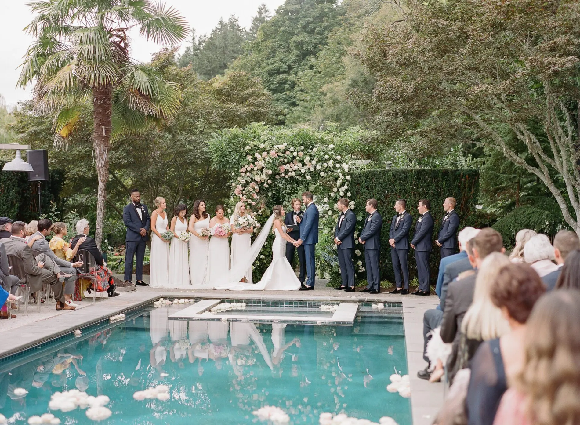 Seattle Catering A wedding ceremony by a pool at a private home, with the couple holding hands, an officiant speaking, bridesmaids in white dresses, groomsmen in dark suits, and guests seated. Floral arrangements decorate the scene. For more inspiration, consider these Private Home Wedding Tips to create your perfect day.