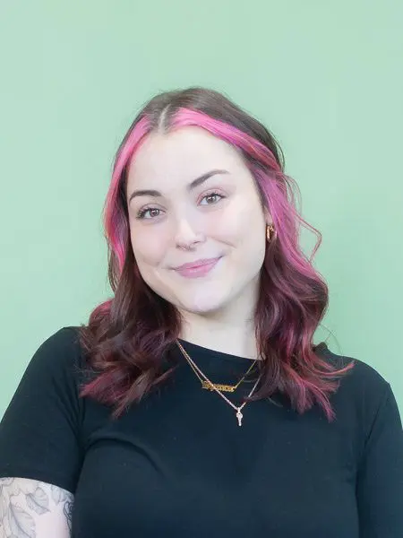 Seattle Catering A person with pink highlights in their hair, wearing a black top and a necklace, stands in front of a green background.