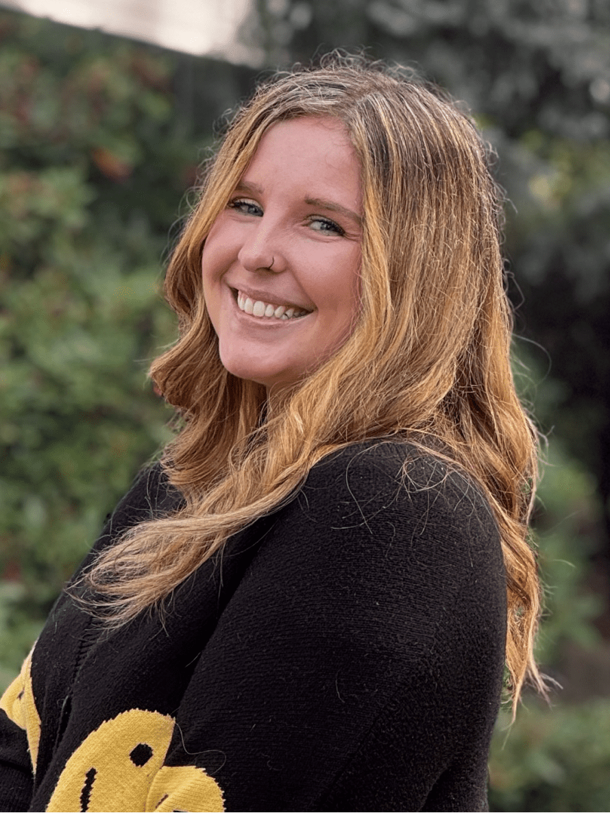 Seattle Catering A person with long blonde hair is smiling at the camera while wearing a black sweater with yellow smiley faces. The background is blurred green foliage.