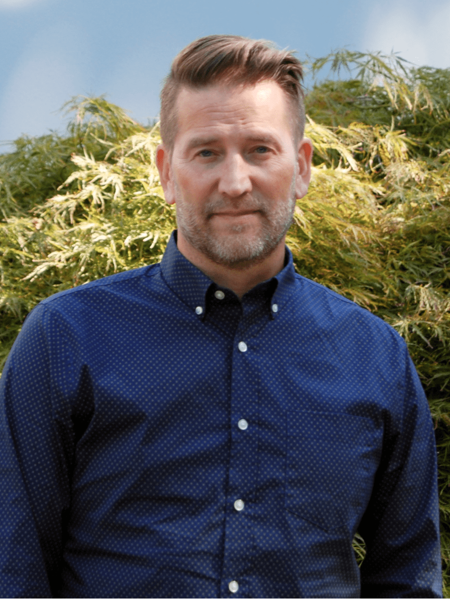 Seattle Catering A man in a blue shirt stands outdoors in front of a background of green foliage, facing the camera.