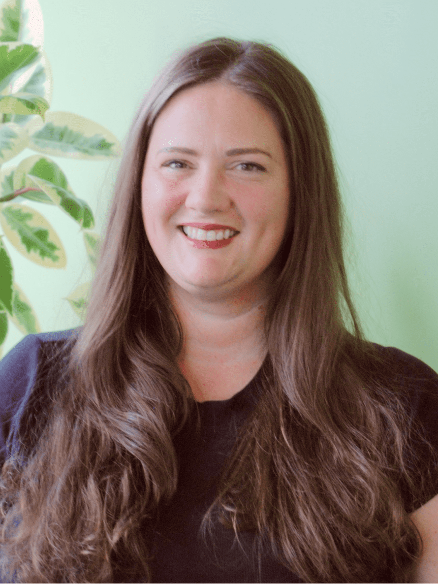 Seattle Catering A woman with long brown hair, smiling, stands in front of a light green wall. She is wearing a black shirt, and a leafy plant is partially visible in the background.