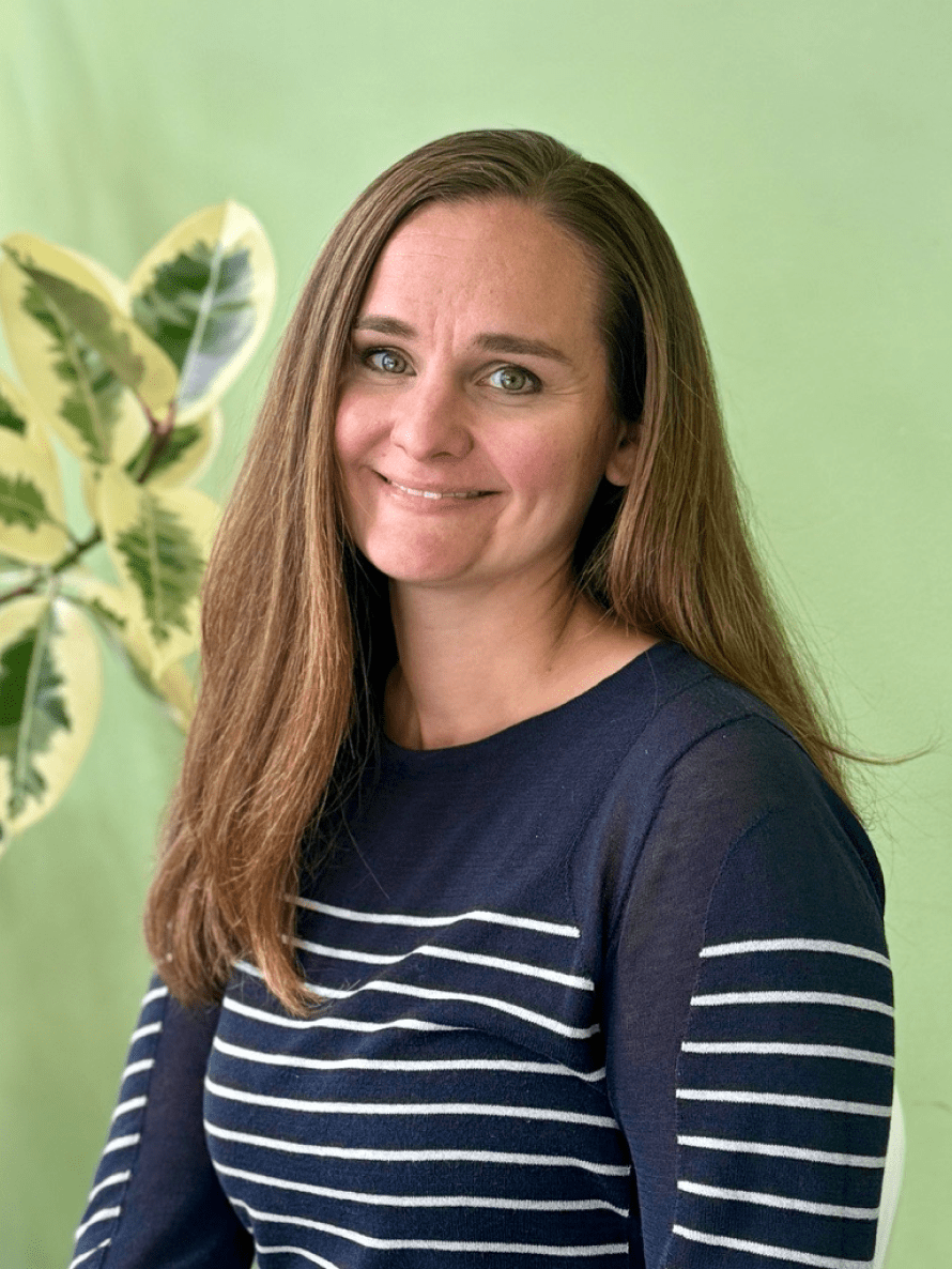 Seattle Catering A woman with long brown hair wearing a navy-blue and white striped shirt smiles in front of a green background with a leafy plant.