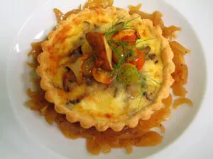 Seattle Catering A small tart filled with a creamy mixture, garnished with mushrooms, herbs, and cherry tomatoes, placed on a bed of caramelized onions.