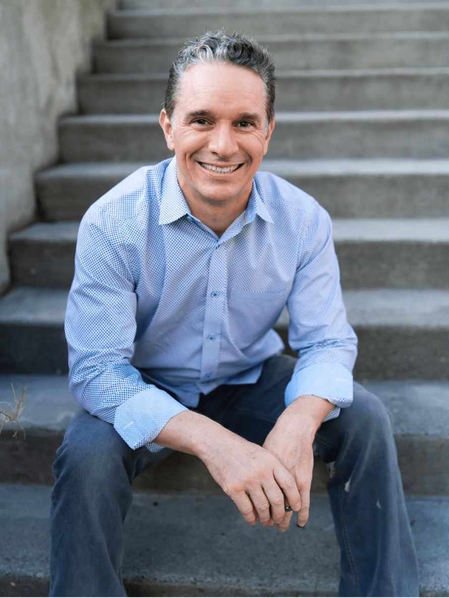 Seattle Catering A man wearing a light blue dress shirt and jeans sits on outdoor concrete steps, smiling at the camera.