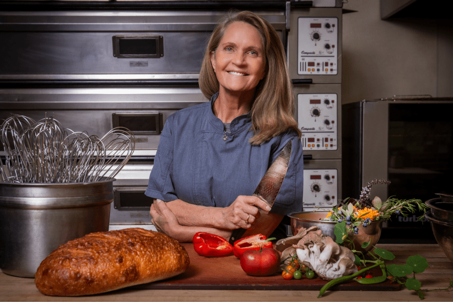 Seattle Catering A woman in a kitchen holding a knife, surrounded by bread, peppers, mushrooms, tomatoes, and fresh herbs. Industrial kitchen equipment in the background.