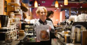 Seattle Catering A woman in a cafe holds up a cookbook titled "For the Love of the South" while smiling. Coffee equipment and cups are visible in the background.