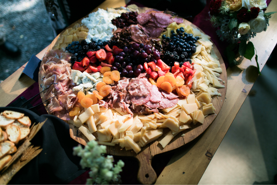 Seattle Catering A wooden board displaying a charcuterie spread with various cheeses, meats, sliced bread, and fruits like strawberries and grapes, accompanied by floral decorations.