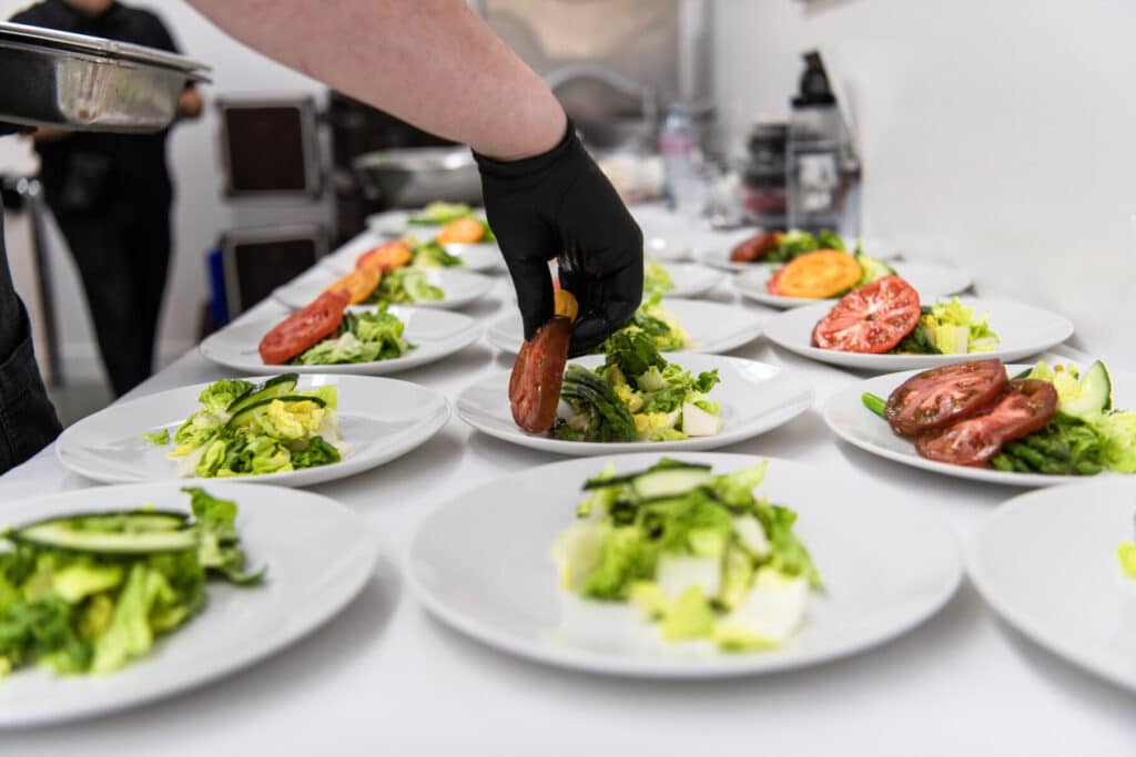 A person wearing a black glove arranges tomato slices on plates of salad lined up on a counter, showcasing the precision and care of wedding catering in Seattle.