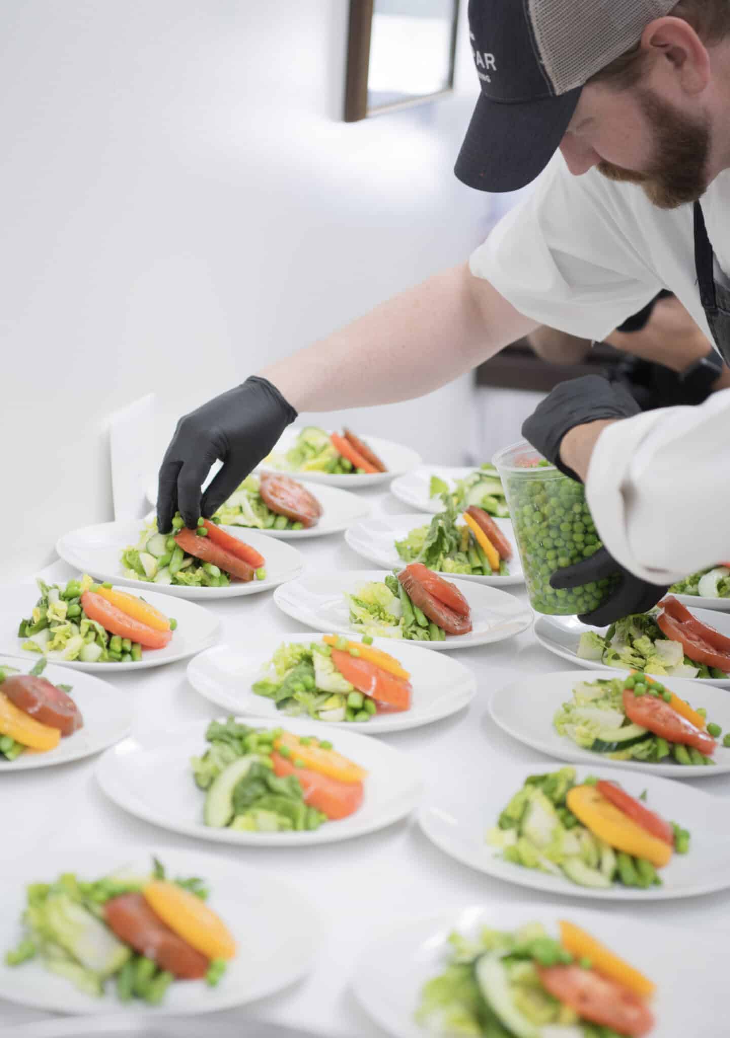 A person wearing gloves arranges green peas onto plates of assorted vegetables in a bright kitchen setting, preparing for elegant wedding catering Seattle style.