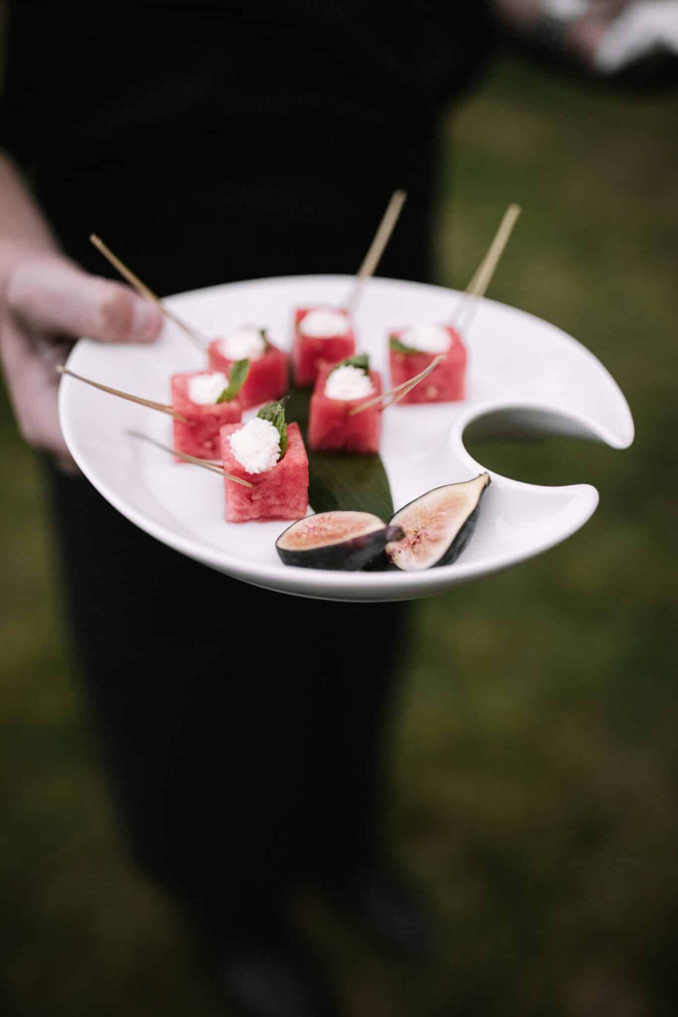 A person holds a white plate with skewered watermelon cubes topped with cheese and mint, alongside two halved figs—an elegant appetizer perfect for wedding catering Seattle style.