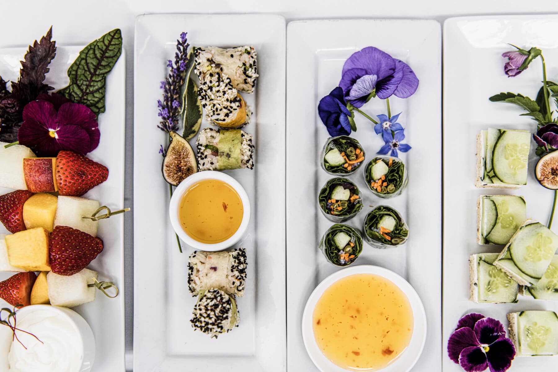 Assorted platters of fruit skewers, sushi rolls, vegetable wraps, and cucumber sandwiches with dipping sauces, garnished with edible flowers and arranged on white plates—perfect for elegant wedding catering in Seattle.