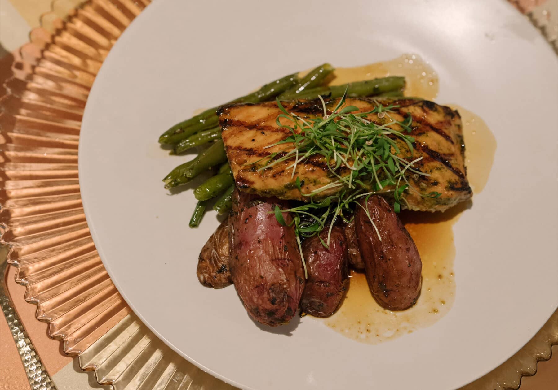 Grilled fish fillet with microgreens, served with green beans and roasted fingerling potatoes on a white plate.