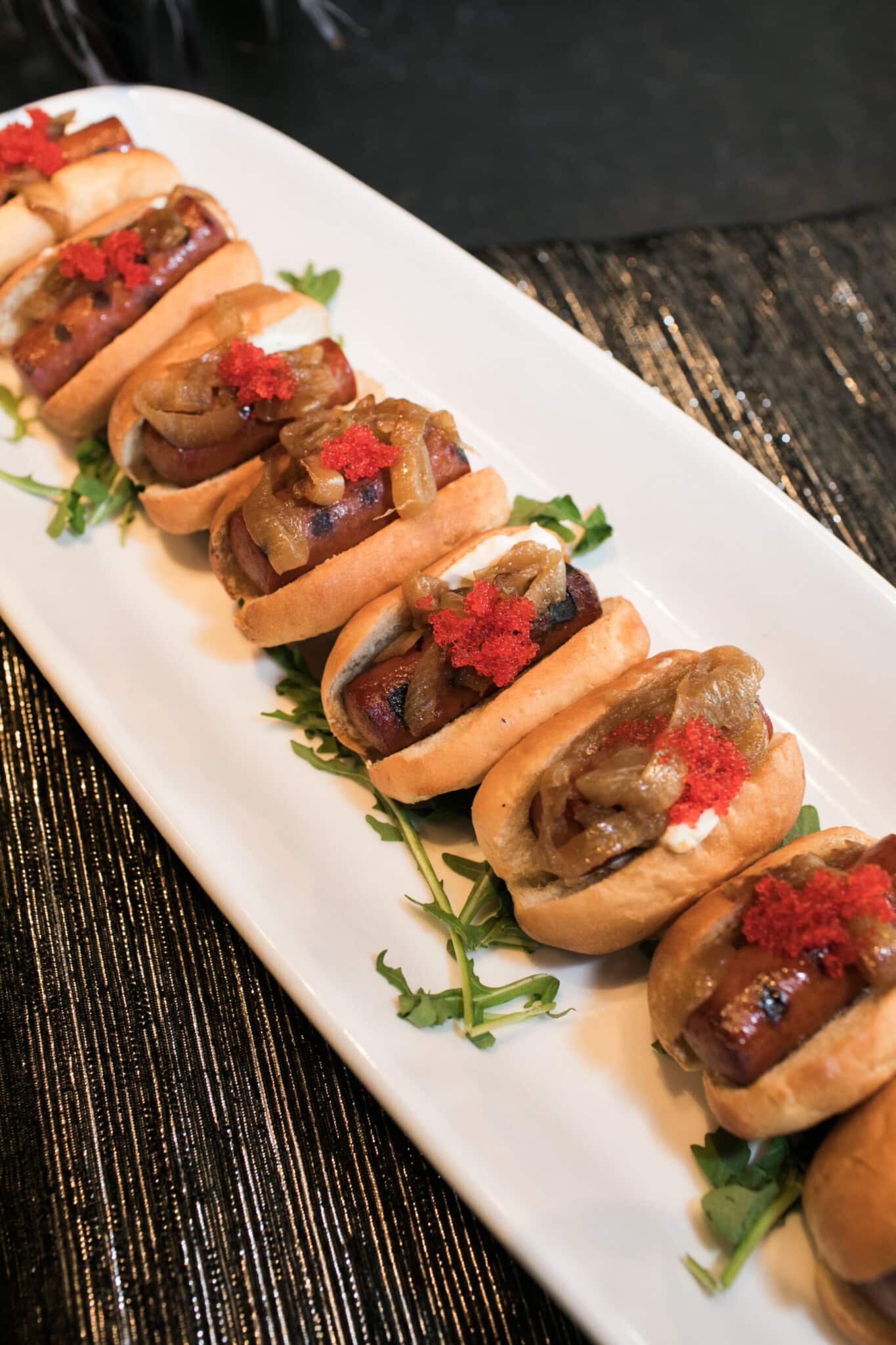 A row of mini sandwiches with grilled meat, caramelized onions, cream, and red garnish, served on a white platter with greens—perfect for wedding and event catering.