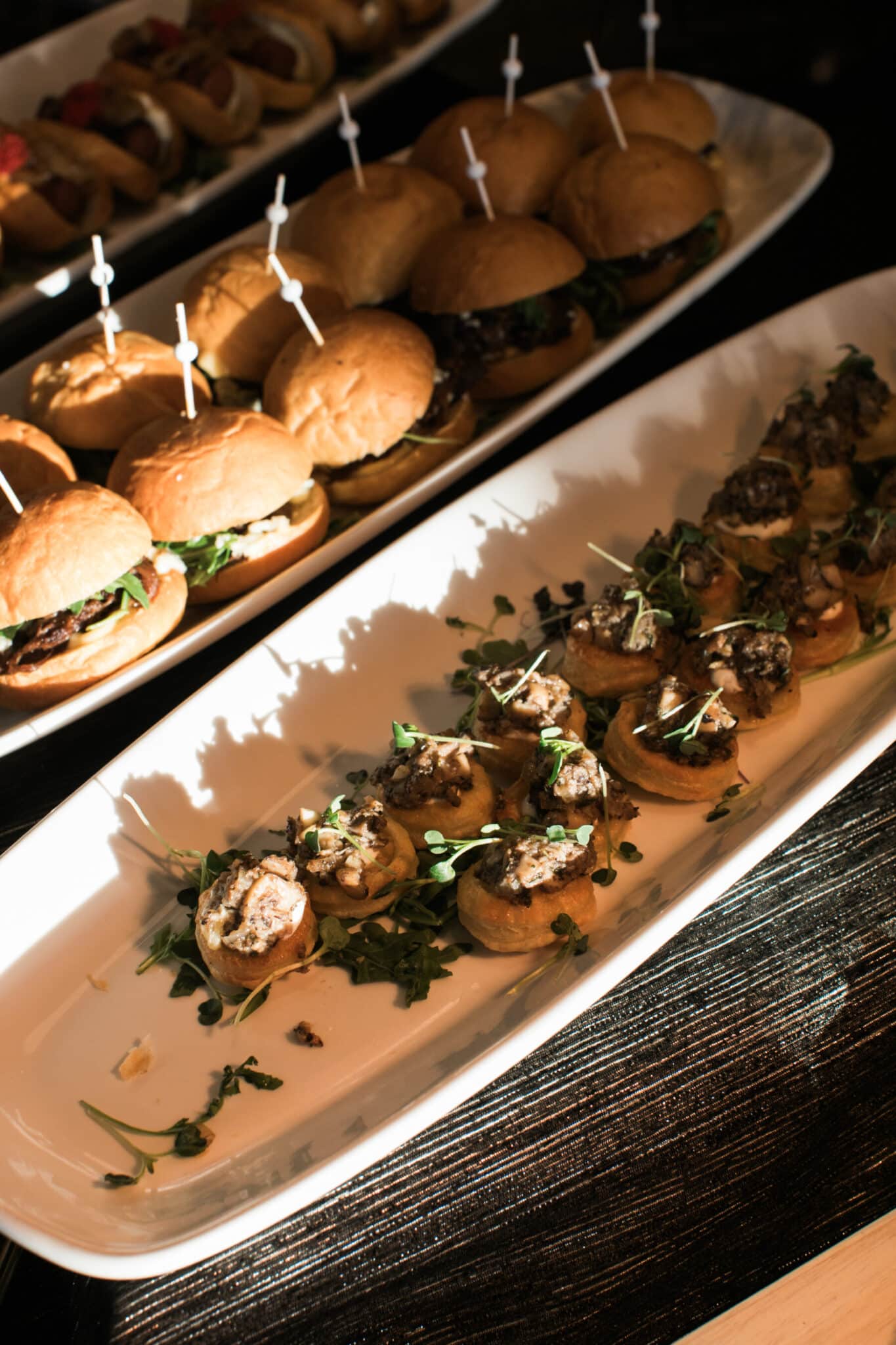 Two platters of appetizers, perfect for wedding and event catering: one with mini sandwiches secured with toothpicks, the other featuring small toasted rounds topped with a savory mixture and fresh greens.