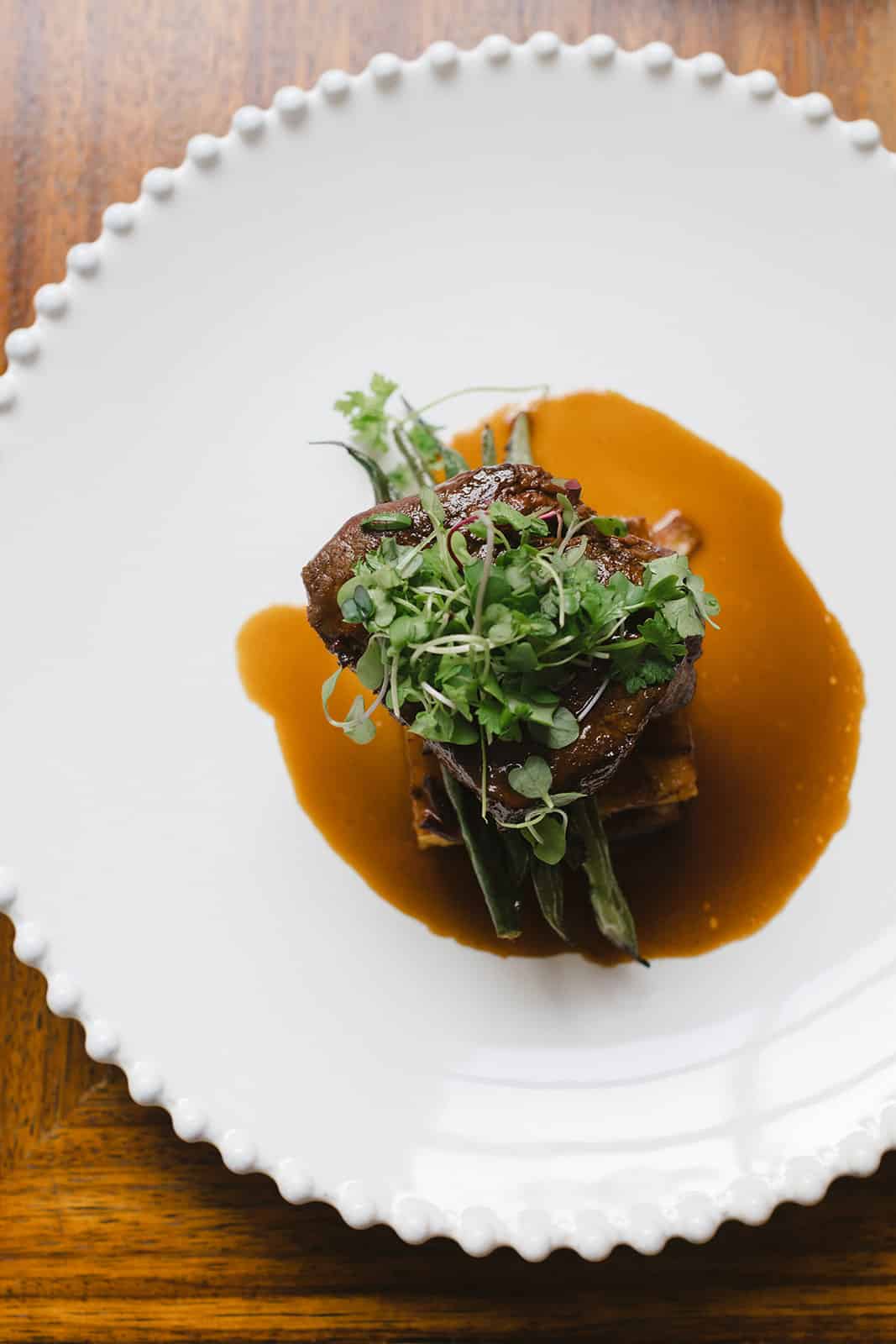 Plated gourmet dish featuring a piece of meat topped with microgreens, served on vegetables with brown sauce on a white plate—perfect for refined wedding and event catering.