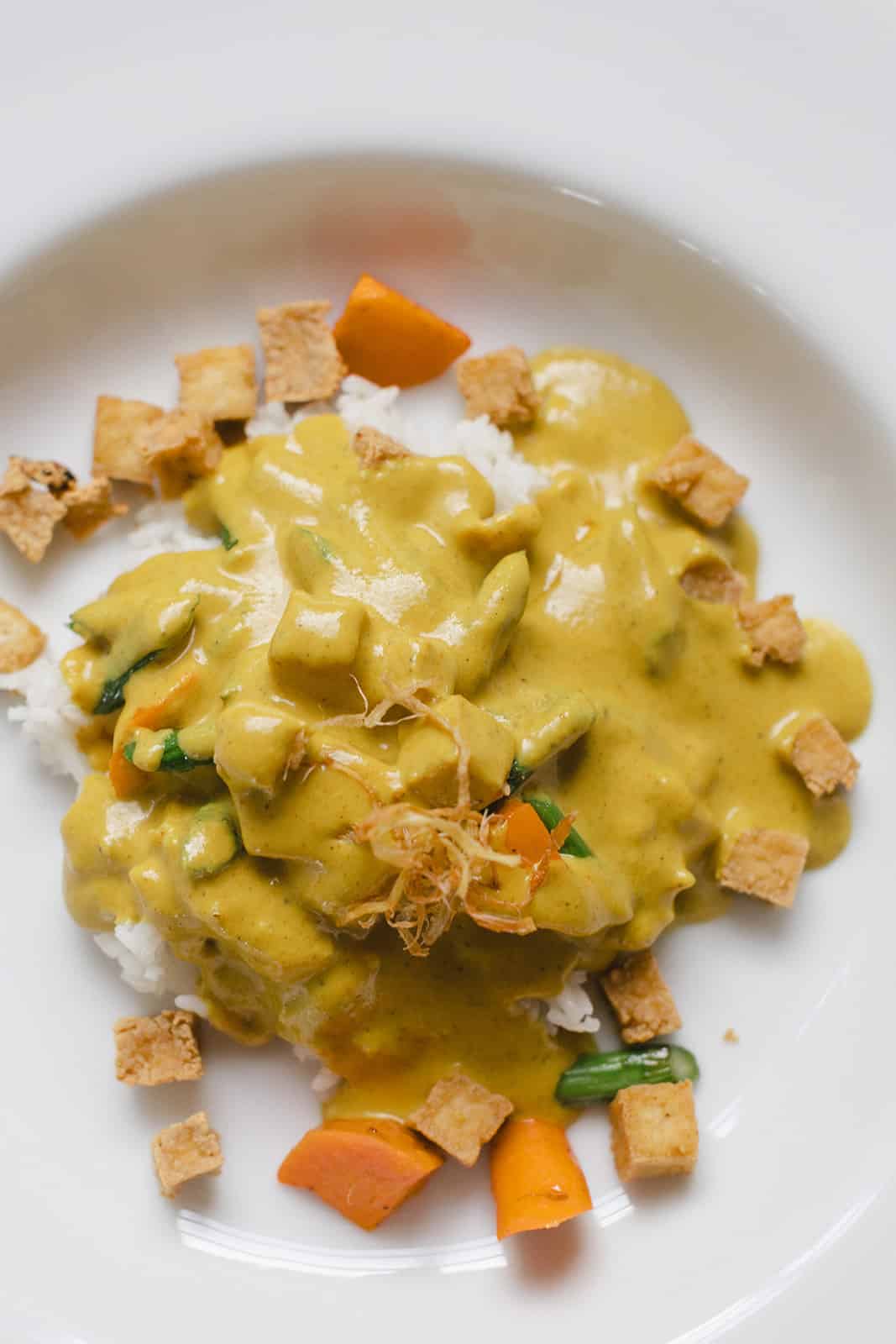 A plate of white rice topped with yellow curry sauce, green beans, orange vegetables, and garnished with crispy tofu cubes and fried shallots—perfect for wedding and event catering.