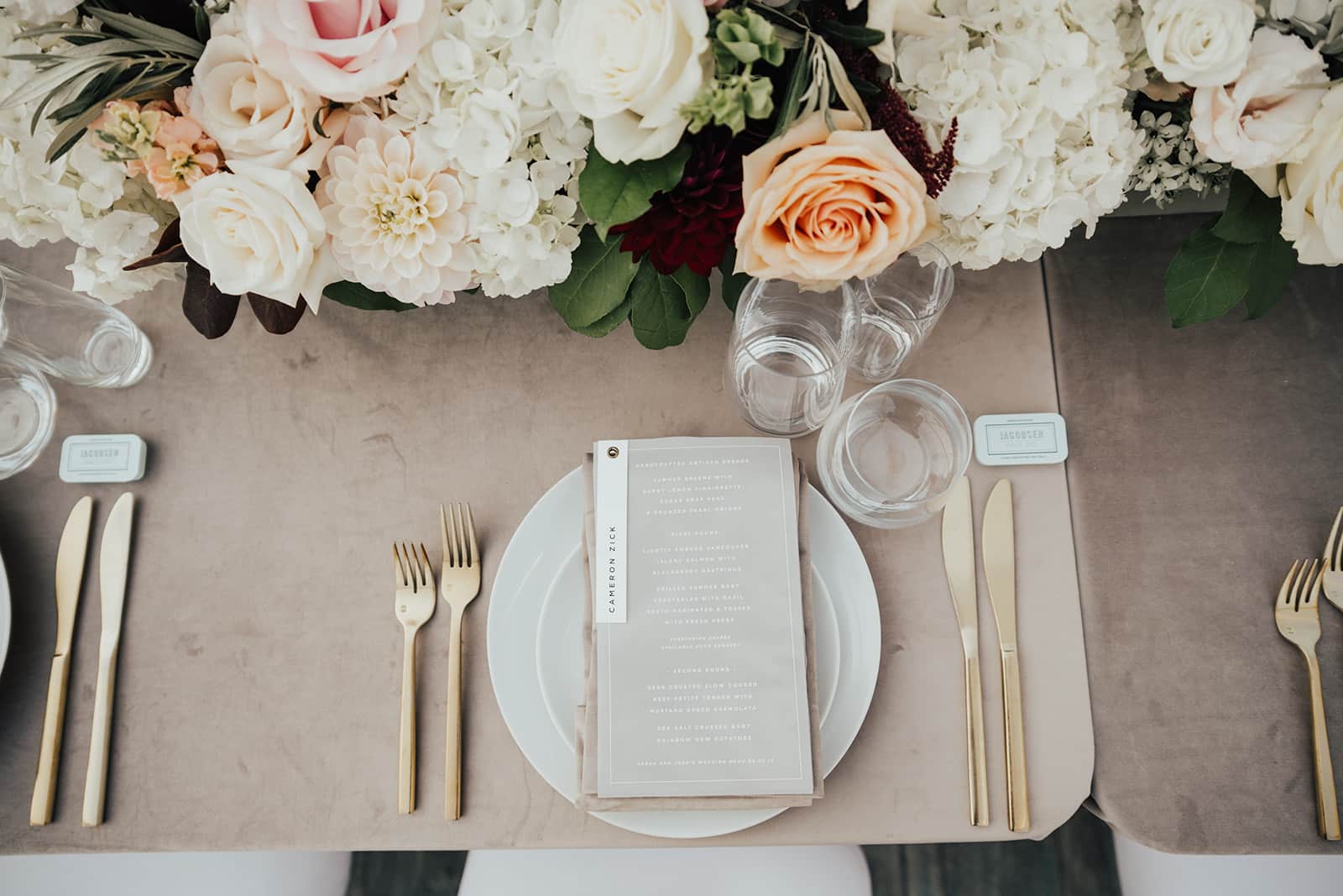 A formal place setting with gold cutlery, a white plate, a printed menu, two glasses, and elegant white and blush blooms on a beige tablecloth—perfect inspiration for wedding catering Seattle couples will love.