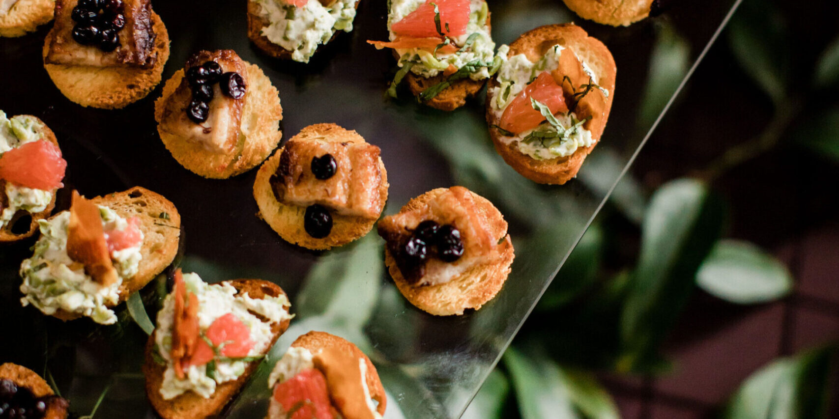 Assorted bite-sized appetizers with toppings like fish, berries, and avocado on round toasted bread, beautifully arranged on a glass surface—perfect for elegant wedding catering in Seattle.