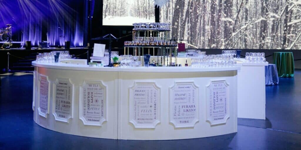 A circular white bar with tiered glass displays and menus is set up in an event venue with winter-themed decor and a forest backdrop, perfect for those seeking top event vendors in Seattle.