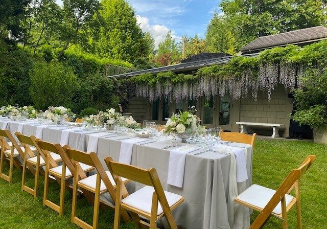 Outdoor dining table set with white linens, floral centerpieces, and wooden chairs on a lawn, perfect for Catering Services in Seattle, with a vine-covered building and trees in the background.