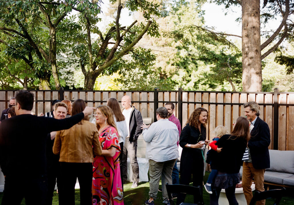 A group of people socializes in a backyard with trees and a wooden fence, some standing and some sitting on outdoor furniture, enjoying delicious wedding and event catering.
