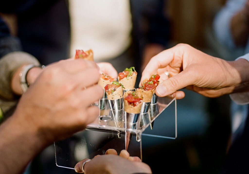 Several hands reach for bite-sized appetizers served in small cones on a transparent tray, showcasing elegant Catering Services in Seattle.