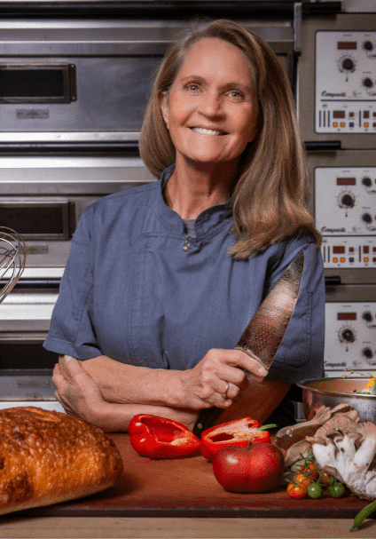 A woman in a chef's coat stands in a kitchen, holding a knife, with bread, vegetables, and baking equipment on the counter—a glimpse behind the scenes at About Lisa Dupar Catering Seattle.