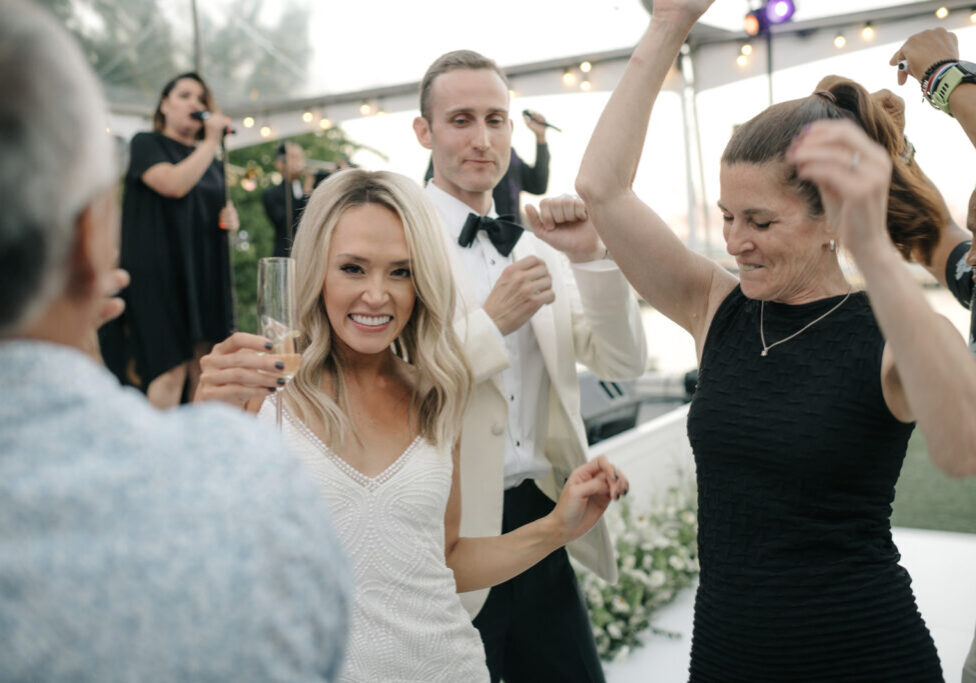 A group of people, including a woman holding a champagne glass, dance and celebrate at a formal event with live music and exquisite wedding and event catering in the background.