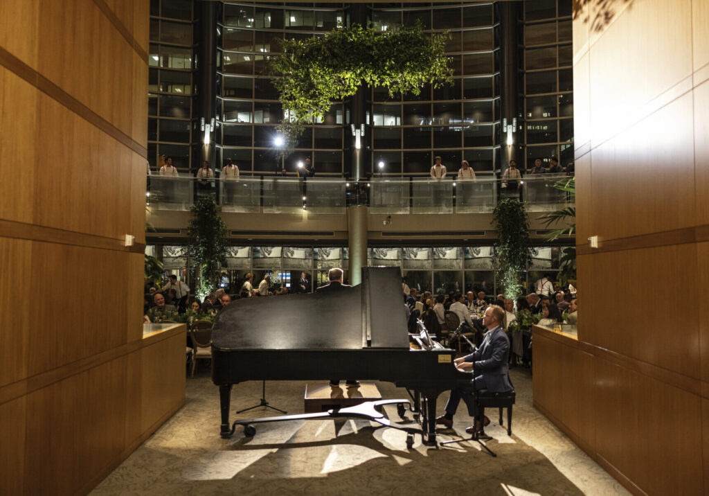 A pianist performs on a grand piano in a spacious, modern atrium, where guests enjoy the music alongside top Catering Services in Seattle, with an audience seated below and people standing on the balcony above.