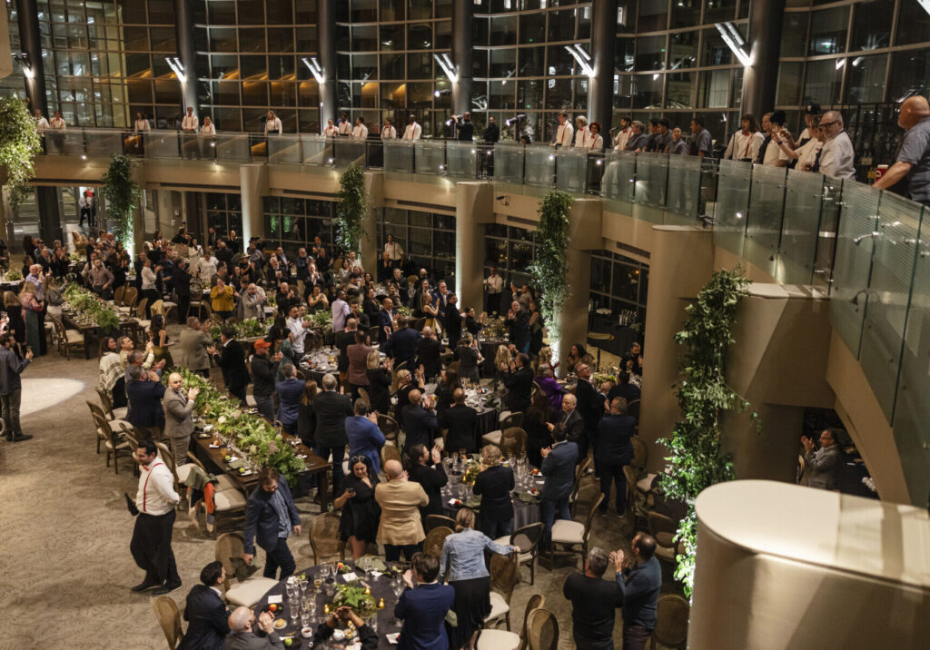 Large group of people attend a formal event in a spacious, modern venue with tables, greenery, and a mezzanine level; many are standing and mingling while enjoying wedding and event catering.