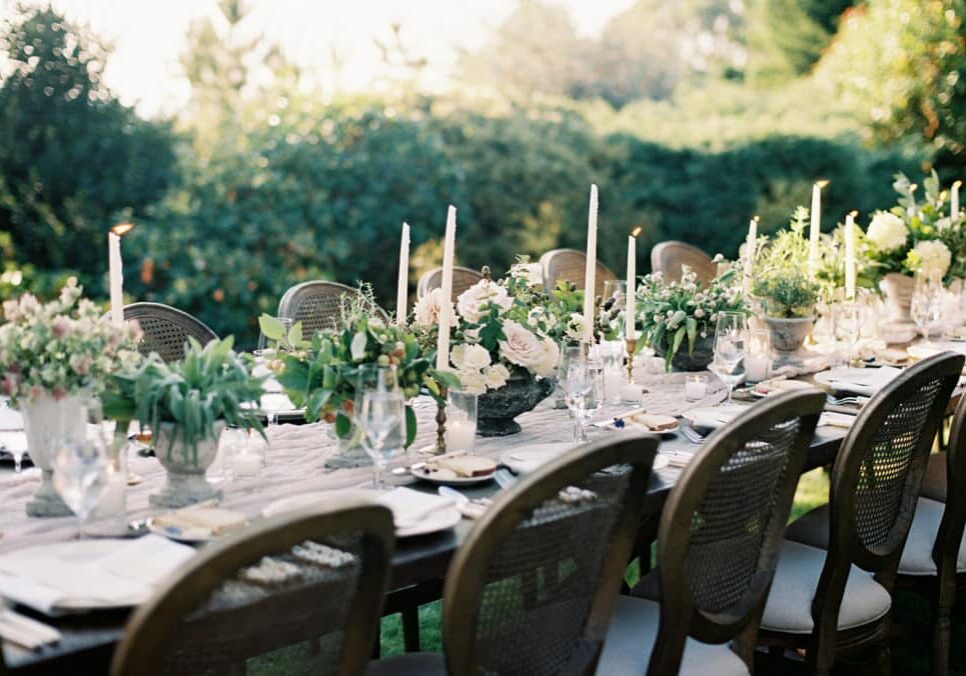 A long outdoor dining table, perfect for wedding catering in Seattle, is set with white candles, floral centerpieces, glassware, and plates, surrounded by wooden chairs in a picturesque garden setting.