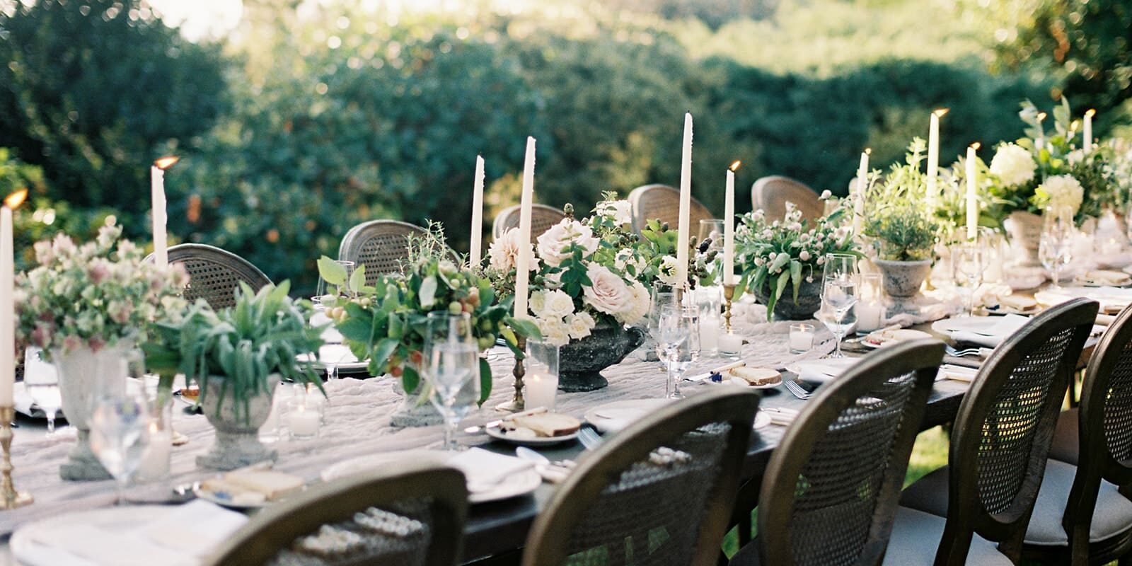A long outdoor dining table, perfect for wedding catering in Seattle, is set with white candles, floral centerpieces, glassware, and plates, surrounded by wooden chairs in a picturesque garden setting.