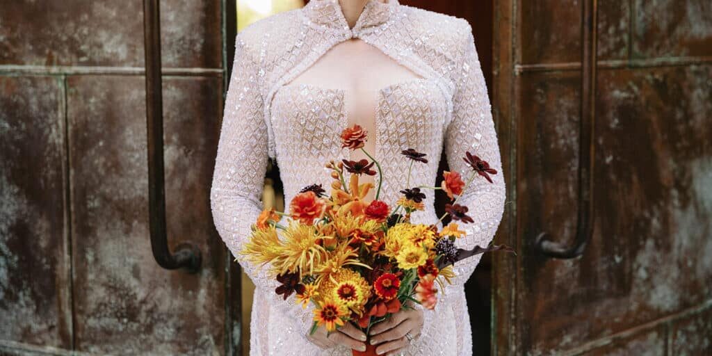 A person wearing an embellished light-colored dress holds a bouquet of yellow, orange, and red flowers, standing in front of large metal doors—perfect inspiration for event vendors in Seattle.