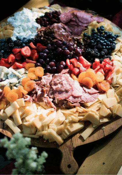 Seattle Catering A wooden board displaying a charcuterie spread with various cheeses, meats, sliced bread, and fruits like strawberries and grapes, accompanied by floral decorations.