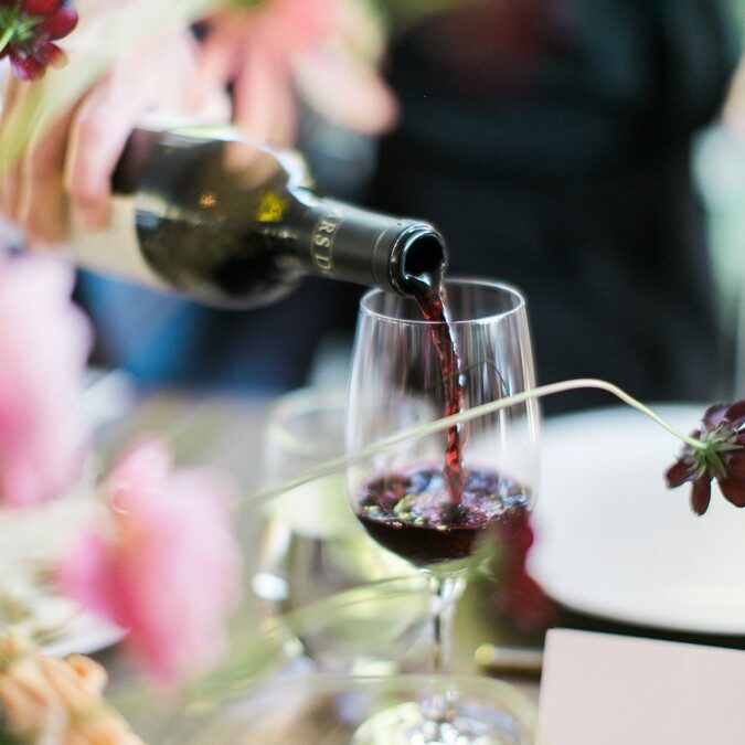 A person pours red wine from a bottle into a glass on a table set for a meal, with flowers and tableware visible—perfectly capturing the elegance of catering for fundraisers.