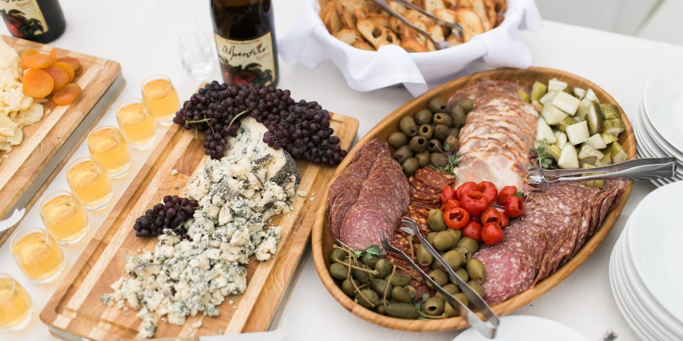 A spread of assorted cheeses, sliced meats, olives, peppers, grapes, bread, and bottles of wine arranged on a white tablecloth—a perfect choice for elegant wedding catering in Seattle.