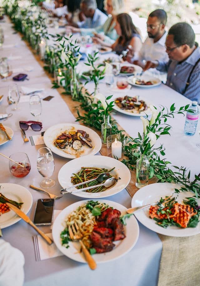 A long table set with plates of food, drinks, and greenery centerpieces creates a warm scene where guests enjoy a meal together—perfect inspiration for wedding catering Seattle style.
